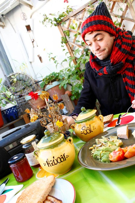 © Johanna Kolb Mann mit Schal und Kappe sitzt am reich gedeckten Frühstückstisch und hört Radio.